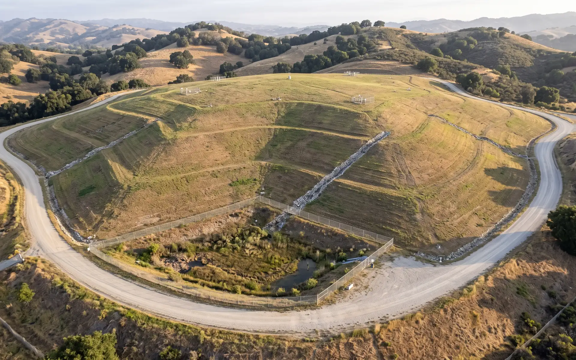 A closed, capped landfill with grass cover, terraced benches, gas well heads and a perimeter road